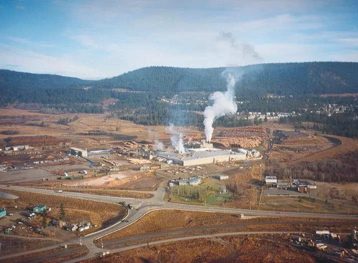 Aerial view of a large industrial plant with smoke rising from chimneys, surrounded by a vast landscape of hills and roads.
