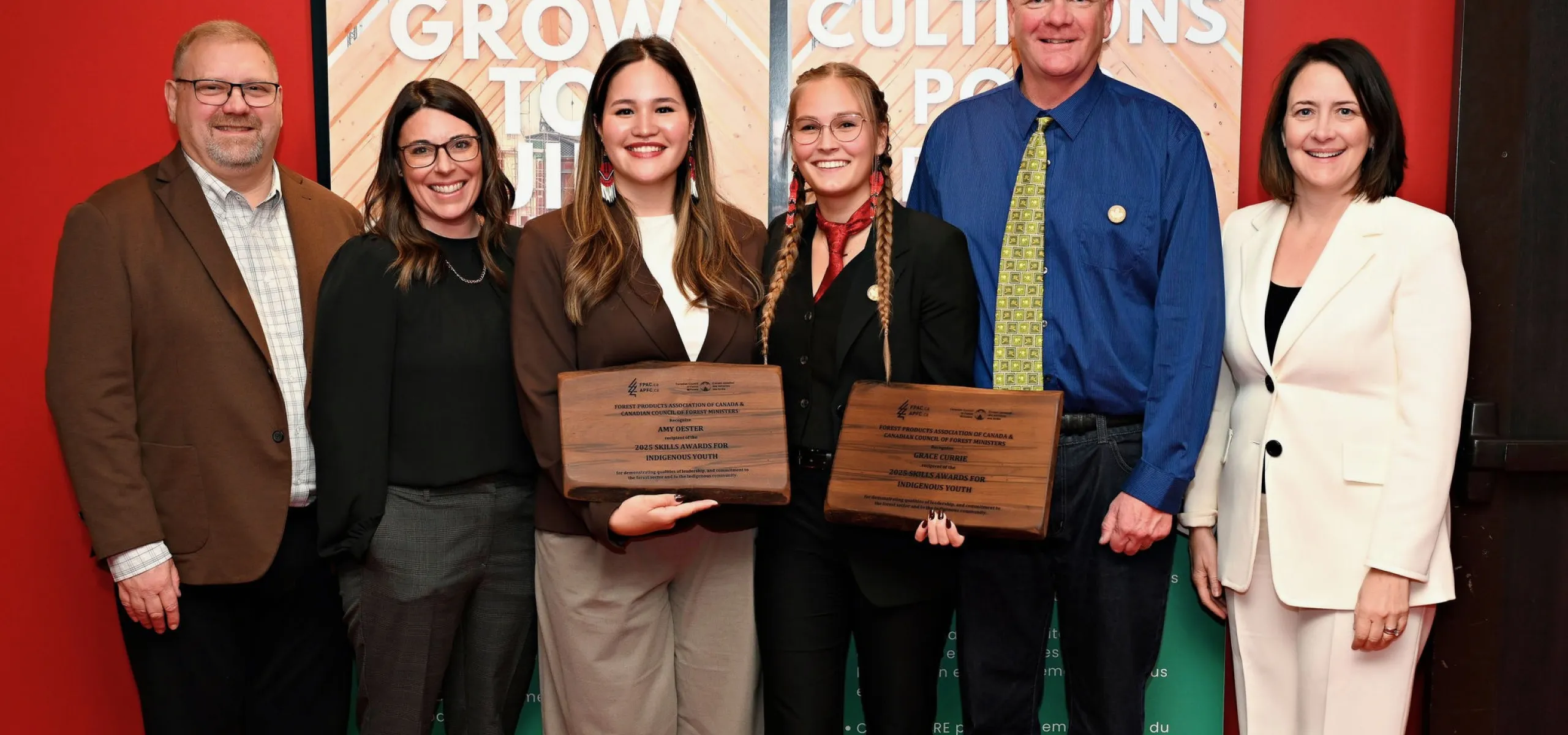 Six people stand indoors, with two women in the center holding award plaques. They are dressed in business attire and are smiling at the camera. Banners and a red wall are in the background.
