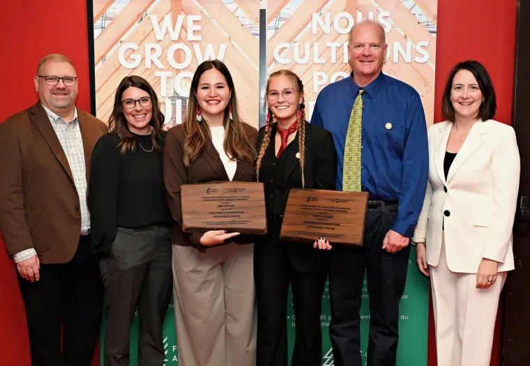 Six people stand indoors, with two women in the center holding award plaques. They are dressed in business attire and are smiling at the camera. Banners and a red wall are in the background.