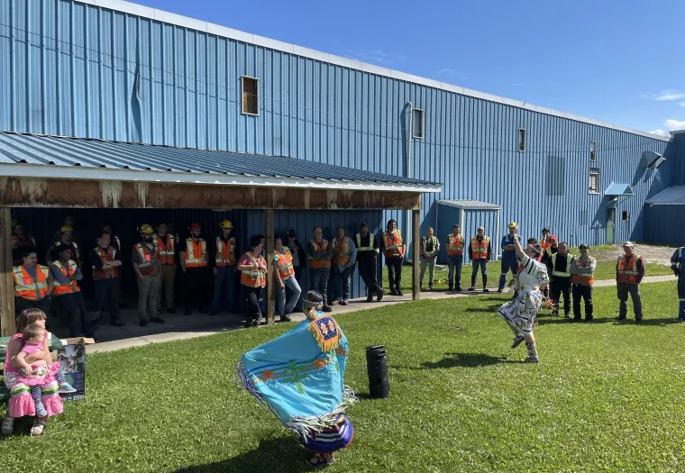 A group of people wearing safety vests stand outside a blue industrial building, observing two dancers in colorful regalia performing on a grassy area. A child sits nearby watching the performance.