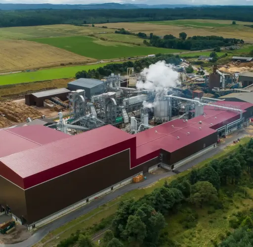 Aerial view of an industrial facility with red roofs emitting steam, surrounded by trees, fields, and stacks of logs in a rural landscape.
